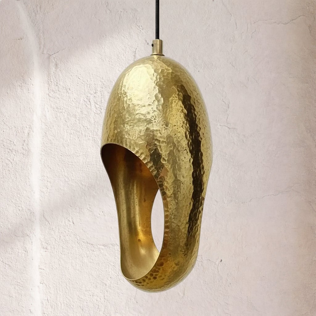Hammered brass pendant lamp with oval side cutout, reflective dimpled surface, suspended from black cord, interior unlit
