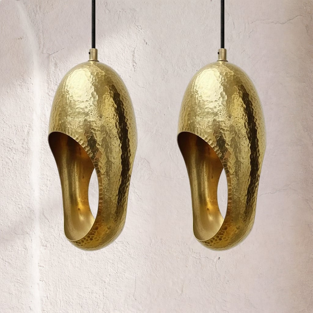 Two hammered brass pendant lamps with oval cutouts, reflective dimpled surfaces, hanging side by side from black cords