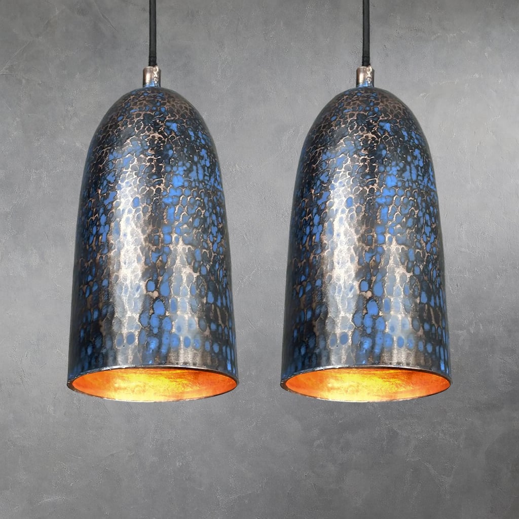 Two hammered copper cylindrical pendant lamps with blue patina textured finish, hanging side by side from black cords, interiors glowing