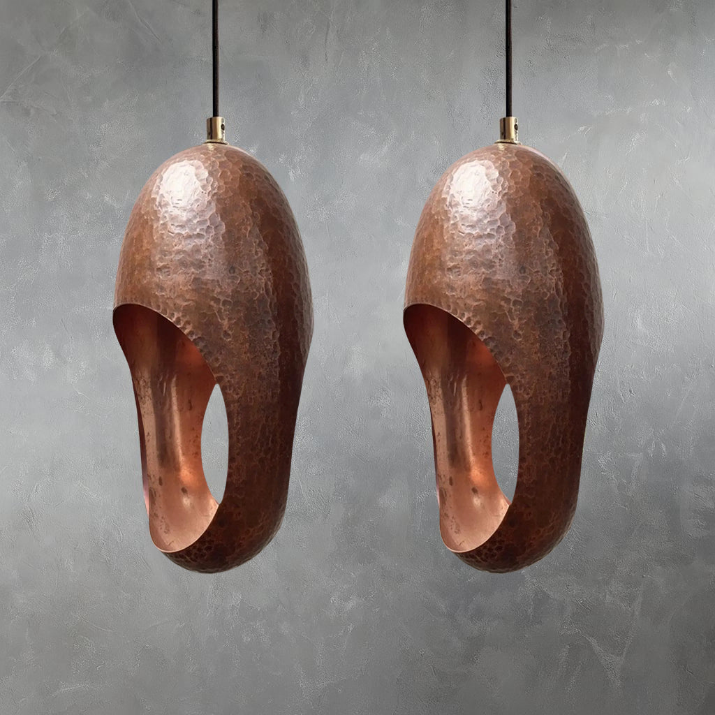 Two copper pendant lights with hand-hammered surface texture and elongated oval openings suspended from cords against gray wall