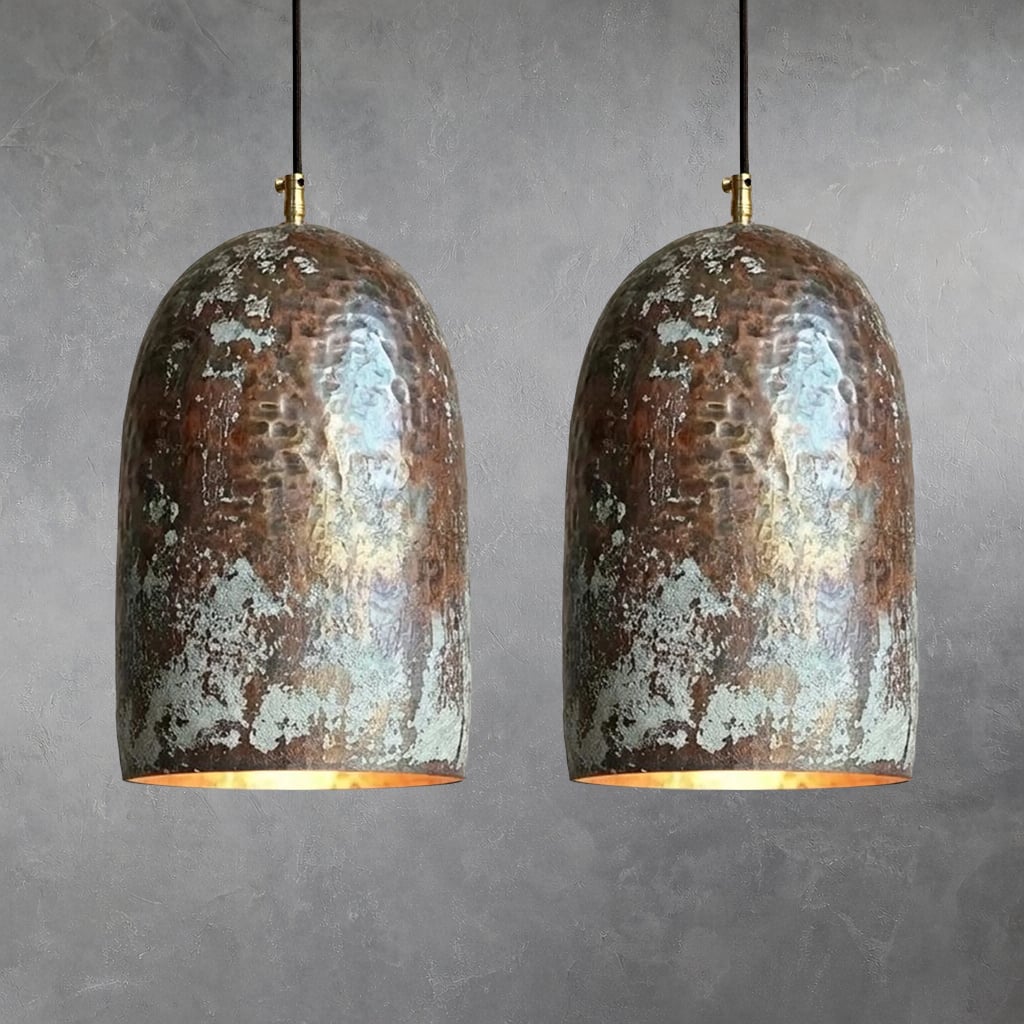 Two hammered copper bell pendant lights with mottled oxidation and verdigris patches, suspended by black cords with brass fittings, warm light emitting from lower openings against neutral wall background