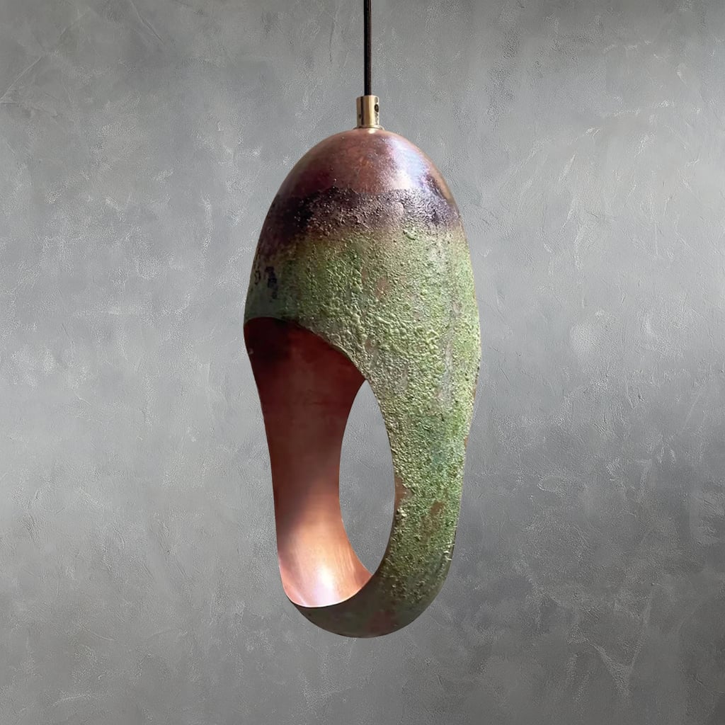 Copper pendant light with hammered surface, green patina oxidation, and elongated oval opening suspended from cord with interior against gray wall