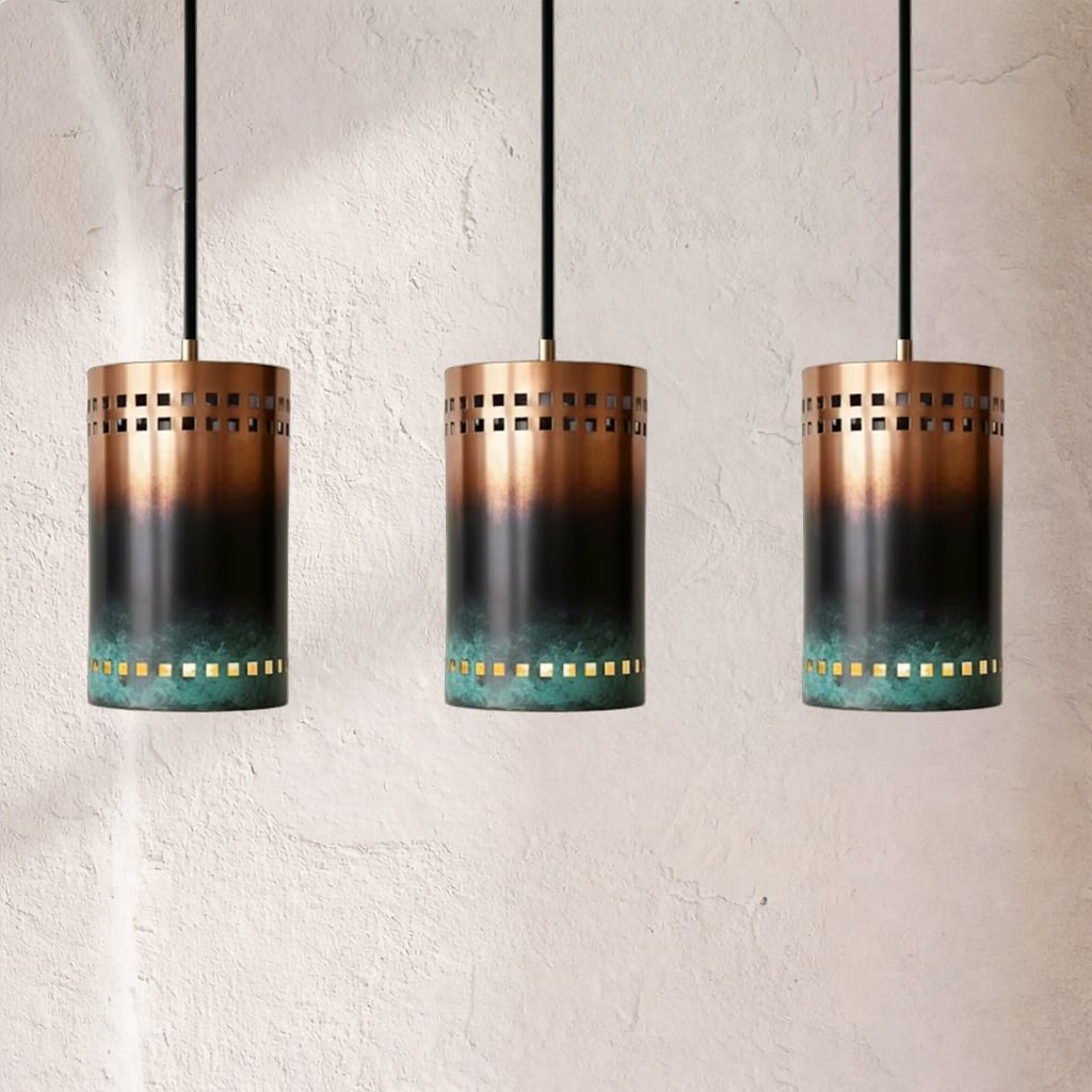 Three cylindrical copper pendants with oxidized gradient finish and green patina base, rows of square perforations around body, suspended by black cords against textured plaster wall