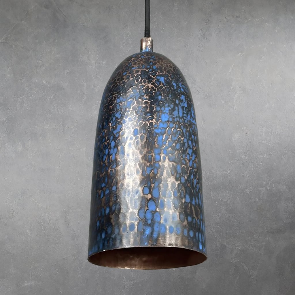 Hammered copper cylindrical pendant lamp with blue patina finish and mottled texture, suspended from black cord, interior unlit