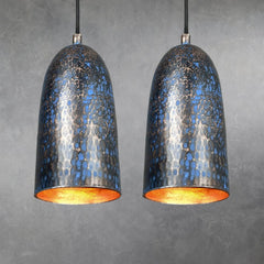 Two hammered copper cylindrical pendant lamps with blue patina textured finish, hanging side by side from black cords, interiors glowing