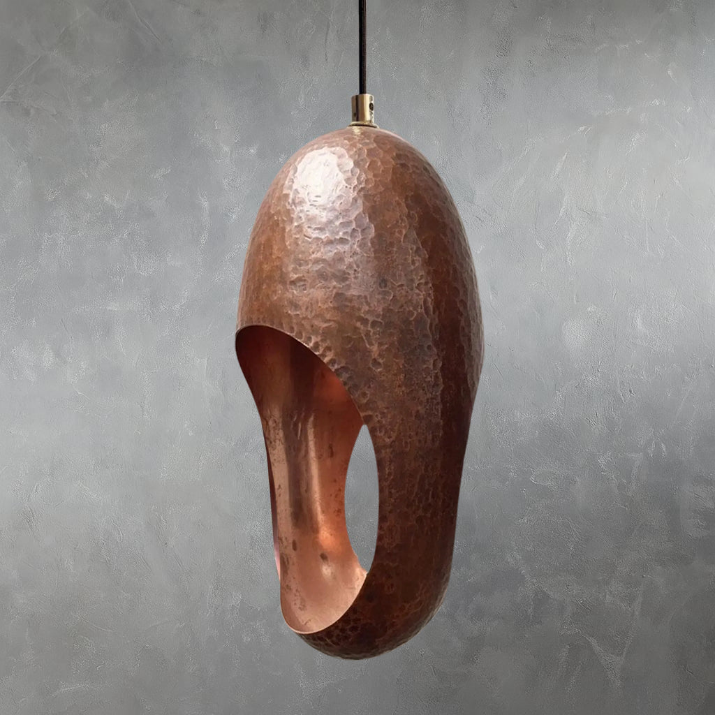 Single copper pendant light with hammered metal texture and elongated oval opening hanging from cord against gray wall