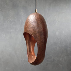 Single copper pendant light with hammered metal texture and elongated oval opening hanging from cord against gray wall