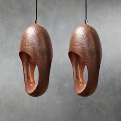 Two copper pendant lights with hand-hammered surface texture and elongated oval openings suspended from cords against gray wall