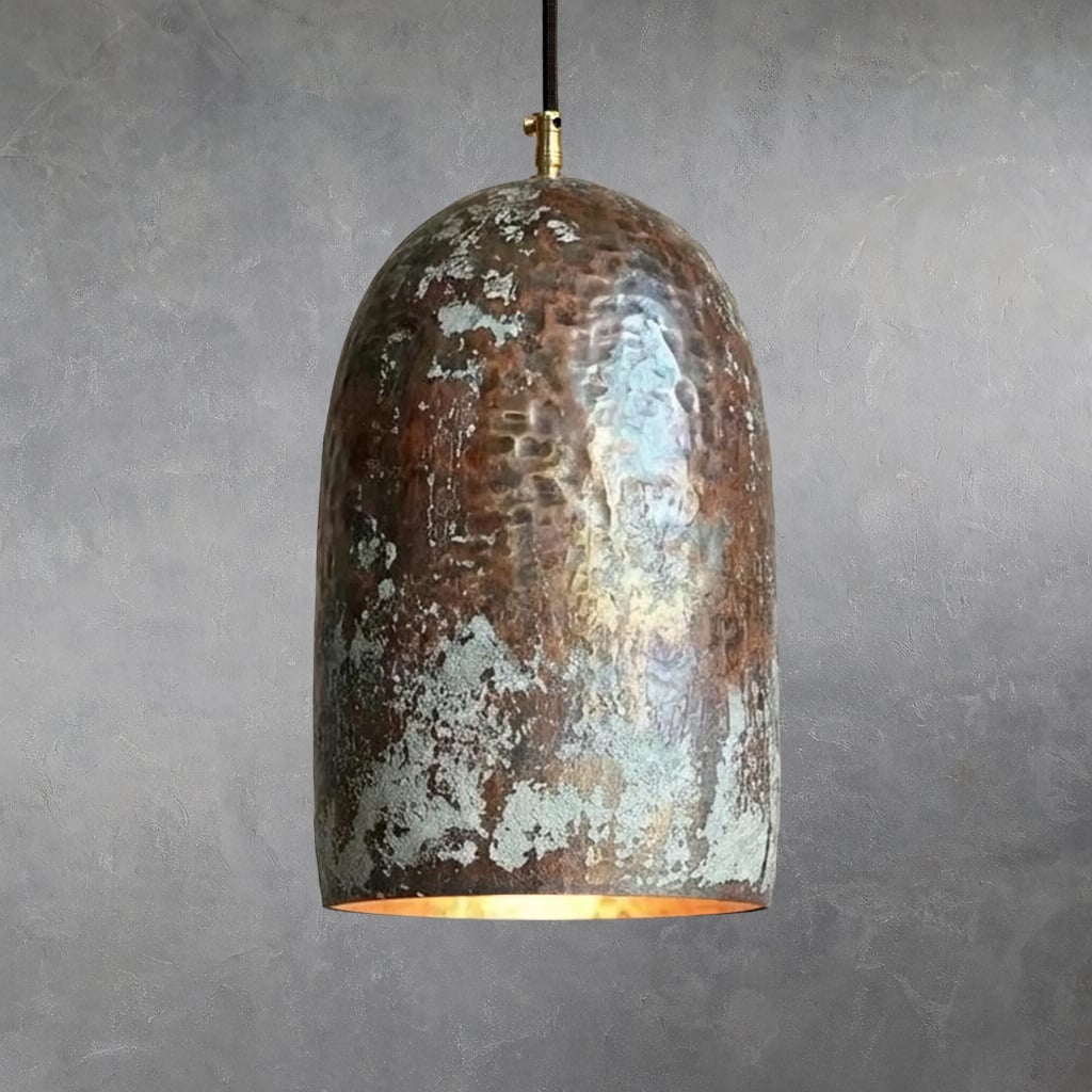 Hammered copper bell pendant with irregular oxidation and verdigris patches, suspended from black cord with brass fitting, warm light visible from lower opening highlighting copper interior