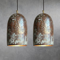 Two hammered copper bell pendant lights with mottled oxidation and verdigris patches, suspended by black cords with brass fittings, warm light emitting from lower openings against neutral wall background