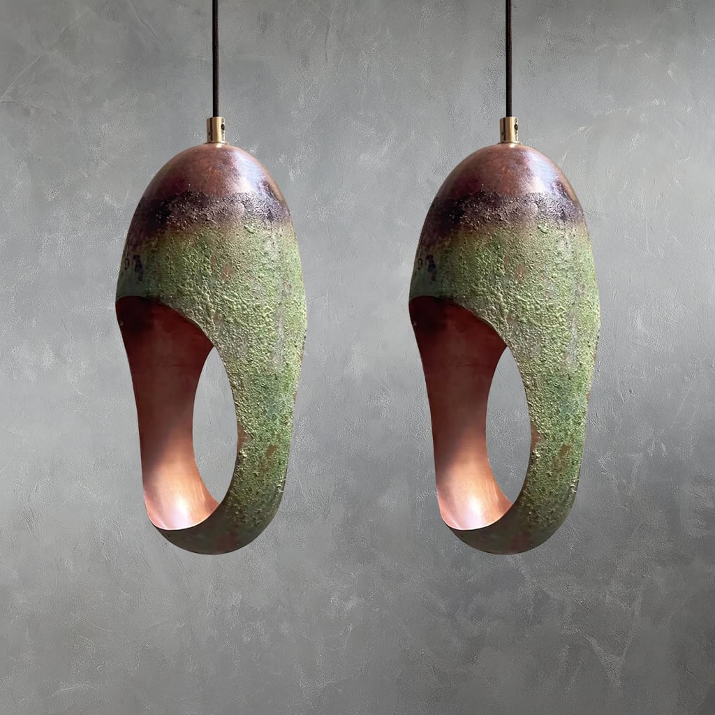 Two copper pendant light with hammered surface, green patina oxidation, and elongated oval opening suspended from cord with interior against gray wall