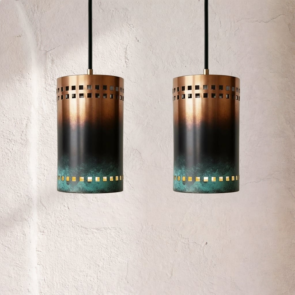 Two cylindrical copper pendants with dark oxidation gradient and green patina base, square perforation rows around upper and lower edges, suspended by black cords against plaster wall