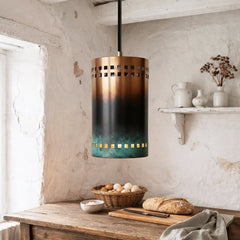 Cylindrical copper pendant with gradient oxidation from warm copper to darkened surface and green patina base, square perforation bands near top and bottom, suspended above wooden table in plaster wall interior