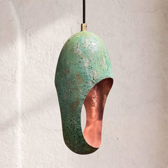 Patinated copper pendant lamp with oxidized green surface and oval side cutout, textured corrosion pattern, hanging from cord against plaster wall